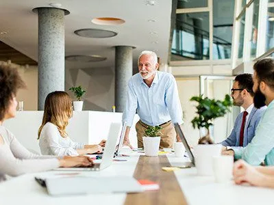 A diverse team sits around a conference table during a meeting, engaged with laptops and notebooks, with plants in the background.